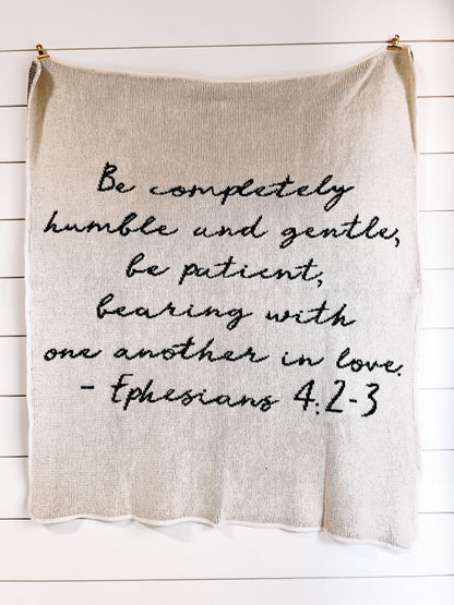Natural Ephesians 4:2-3 blanket with black handwritten scripture, hanging against a white plank wall.