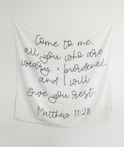 A black and white muslin swaddle blanket with text printed on it hanging on a wall, with a wooden crib in the foreground.