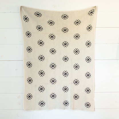 Natural knit blanket with small black snowflakes, shown fully extended against a white plank wall.