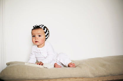 Baby in a white coverall and striped headband seated on a cushion