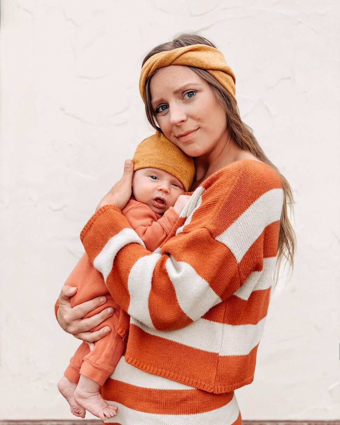 Adult holding a baby wearing a rust-colored thermal zip coverall and mustard knit hat.