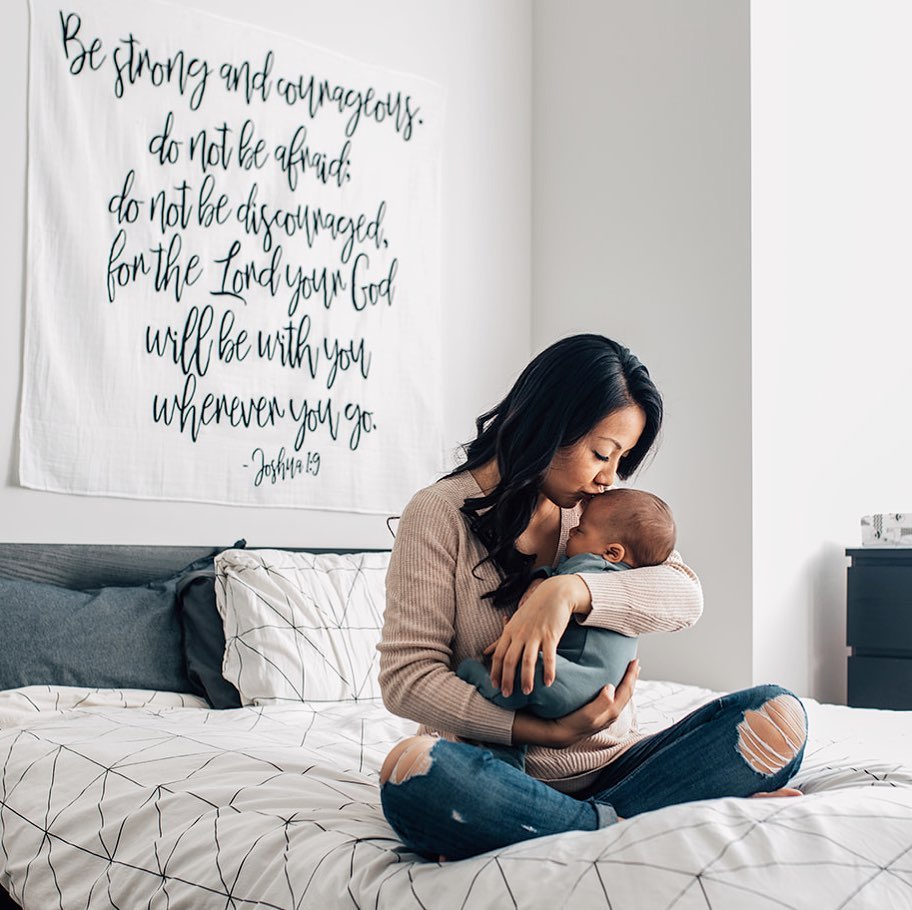 A woman sitting on a bed with a swaddle blanket hanging on the wall behind her. The blanket has black and white geometric patterns and the wall art features text.