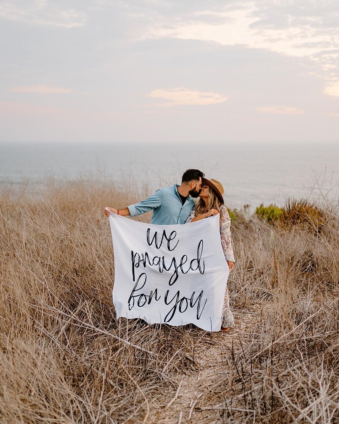 A couple holding a white swaddle blanket with the text 'we prayed for you' written in a script font, standing in a field.
