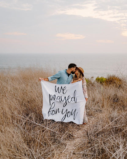 A couple holding a white swaddle blanket with the text 'we prayed for you' written in a script font, standing in a field.